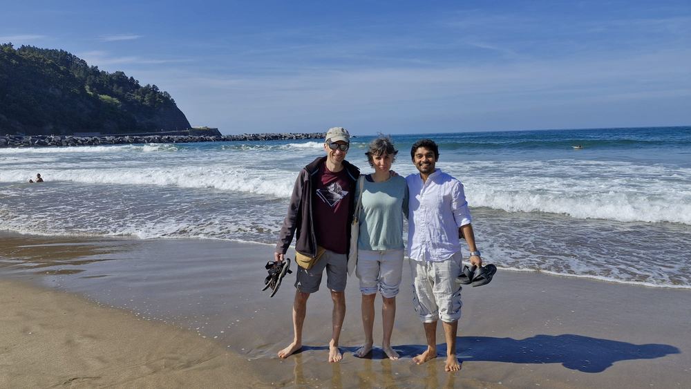 Abhiram barefoot on a sunny Basque beach with two friends, waves and a green headland behind them