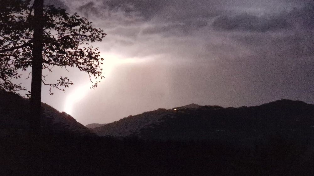 Lightning lighting up the night sky over dark Basque hills, with a tree silhouetted on the left